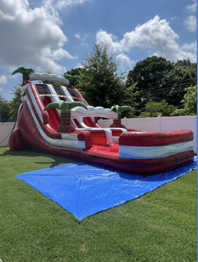 Water Slide Rentals in Naples, FL: red and white tropical-themed dual-lane water slide with palm tree accents, set up on a grass lawn for a backyard party