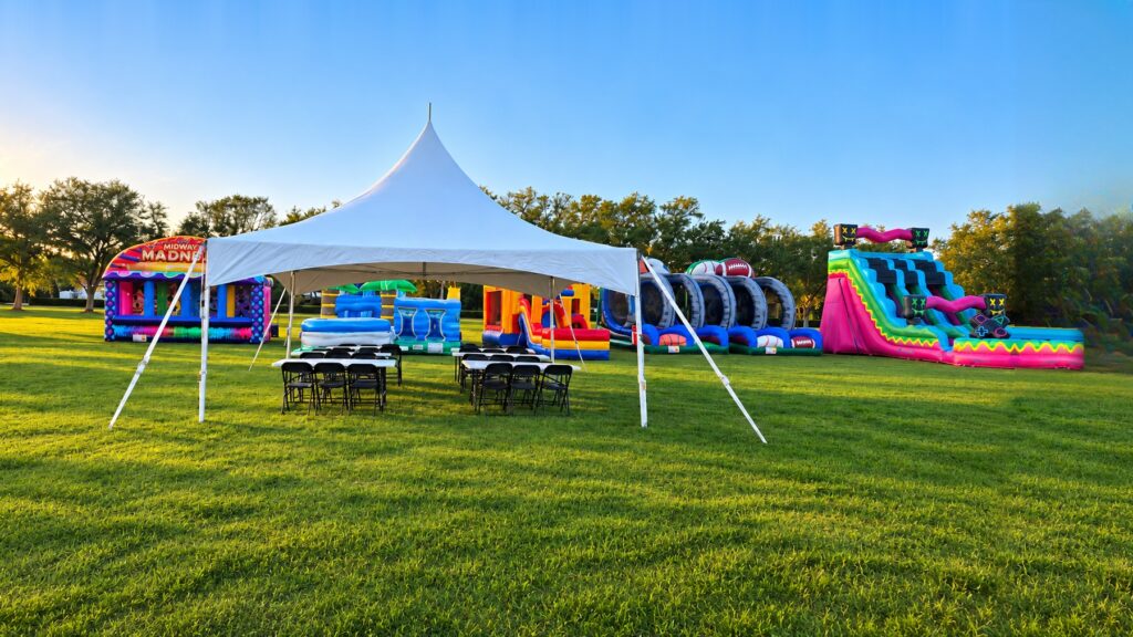 A white frame tent with black folding chairs and tables on a grassy lawn in Fort Myers, FL, with a Midway Madness carnival game, a bounce house, an obstacle course and a pink water slide set up behind it.